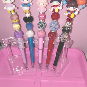 May include: Five decorative pens with character toppers, including a bow-wearing character, Donald Duck, a character with a bow, Mickey Mouse, and Minnie Mouse. Each pen features a different color barrel and decorative beads.