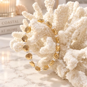 May include: A circular necklace featuring alternating gold-toned metal seashell charms and small white spherical beads. The jewelry piece is arranged in a continuous loop, showcasing a repeating pattern of metallic shell motifs separated by individual white beads.