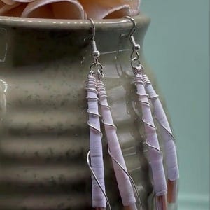 May include: A pair of dangle earrings featuring three pale lavender cylindrical components wrapped in thin silver-toned wire. Each component is capped with a small, translucent, light brown bead at the bottom, suspended from silver-toned hooks.