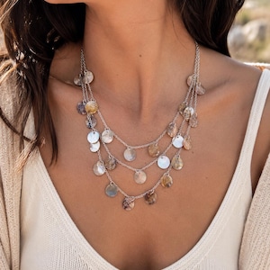 May include: A multi-strand silver-toned necklace featuring three delicate chains adorned with numerous small, circular, iridescent shell-like discs in shades of white, beige, and brown, creating a layered effect around the neck area.