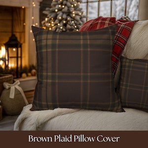 Puede incluir: Funda de cojín a cuadros marrones con un patrón de cuadros clásico en tonos marrones, verdes y beige. La almohada es de forma cuadrada y descansa sobre una superficie blanca y esponjosa. El texto "Brown Plaid Pillow Cover" está en la parte inferior.