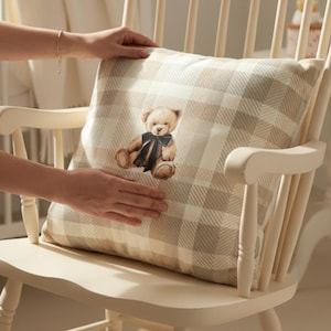 Puede incluir: Una mecedora color crema con un cojín a cuadros con un diseño de oso de peluche. El cojín tiene un patrón a cuadros beige y blanco. El oso de peluche lleva una pajarita azul oscuro. La silla está en un entorno de guardería.