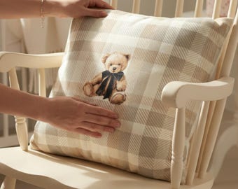 Op de afbeelding: Een crèmekleurige schommelstoel met een geruit kussen met een teddybeerontwerp. Het kussen heeft een beige en wit geblokt patroon. De teddybeer draagt een donkerblauwe vlinderdas. De stoel staat in een kinderkamer.