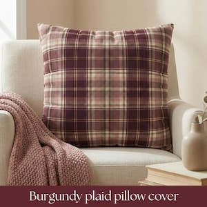 May include: A burgundy plaid pillow cover rests on a cream-colored armchair. The pillow features a classic plaid pattern in shades of burgundy, cream, and tan. A matching knitted throw blanket is draped over the arm of the chair. The text "Burgundy plaid pillow cover" is at the bottom.