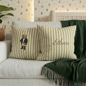May include: Two decorative throw pillows with vertical olive green and white stripes. One pillow features a cartoon bear in a green jacket and khaki pants. The other pillow has the name "Ethan" embroidered in a cursive font. A green blanket is draped over the sofa.