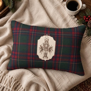 May include: A rectangular throw pillow featuring a dark green, red, and thin gold plaid pattern. Centered on the pillow is a cream-colored scalloped label with a detailed black and white line drawing of a nutcracker surrounded by pine branches.