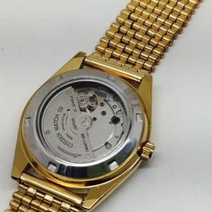 May include: A gold-tone wristwatch with a metallic link bracelet and an exhibition case back. The movement is visible, featuring the text Citizen Watch Co, 21 Jewels, and Japan. The watch has a circular case and a gold-colored crown on the side.