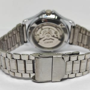 Puede incluir: Reloj de pulsera plateado con una correa de acero inoxidable. La parte trasera del reloj es visible, mostrando el mecanismo interno. La correa se sujeta con un cierre metálico. Un accesorio clásico.