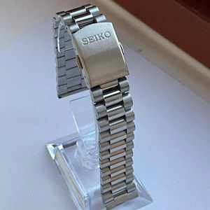 May include: A silver-toned stainless steel watch bracelet featuring a brushed finish and a fold-over clasp. The clasp is engraved with the brand name SEIKO in capital letters. The metal links are arranged in a multi-piece design.