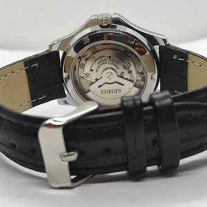 Peut inclure: Gros plan d'une montre Seiko avec un bracelet en cuir noir. La montre a un boîtier argenté et un dos transparent, révélant le mécanisme interne. Le bracelet est fixé avec une boucle argentée.