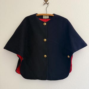 May include: A navy blue cape with a vibrant red lining, featuring a round neckline and three gold-toned metal buttons down the front. The garment has a structured silhouette with additional gold buttons on the sides and a small interior label.