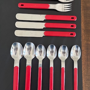 May include: A set of vintage flatware with red handles. The set includes two forks, two knives, and six spoons. The stainless steel utensils have a bright red handle with a hole at the end for hanging. The flatware is arranged on a black surface.