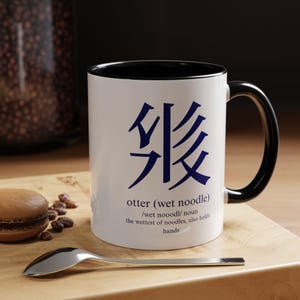 Puede incluir: Una taza de cerámica blanca con interior y asa negros. El frente presenta un texto azul oscuro estilizado que se asemeja a caracteres asiáticos sobre el texto en inglés: otter (wet noodle) /wet nooodl/ noun the wettest of noodles, also holds hands.