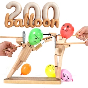 May include: A wooden toy with two figures, each with a balloon head, poised as if about to be pierced by a blade. The text "200 balloon" is above the figures. Several colourful balloons are scattered around the base of the toy.