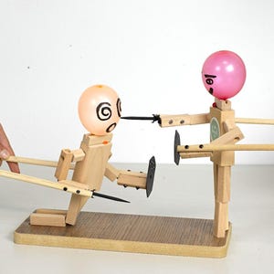 May include: A wooden toy depicting two figures with balloon heads. One figure kneels, facing the other, with a black blade aimed at its face. The other figure has a pink balloon head and holds a blade. The toy is on a wooden base.