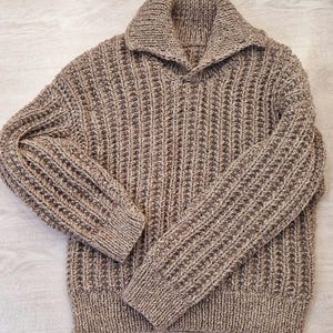 Op de afbeelding: Een bruine, getextureerde gebreide trui met een brede kraag. De trui heeft een ribbelpatroon en lange mouwen. De kleur is een mix van bruin en beige, waardoor een gemêleerd effect ontstaat. De kraag is omgeslagen en de trui lijkt gemaakt van dik, warm materiaal.
