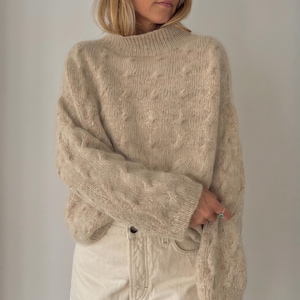 May include: A beige long-sleeve sweater with a mock neck and a textured cable knit pattern. The sweater is paired with off-white denim pants featuring a visible button fly and a silver ring with a small green stone on the finger.