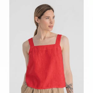 May include: A bright red sleeveless tank top featuring a straight square neckline and wide shoulder straps. The garment is constructed from a textured fabric and has a relaxed, straight-cut silhouette that falls to the waistline.