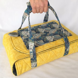 May include: A quilted yellow fabric casserole carrier featuring a dark blue panel with a repeating fan pattern. The carrier includes two matching fabric handles and a silver metal spoon tucked into the side flap, designed for transporting dishes.