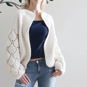 May include: An off-white, long-sleeved, open-front crochet cardigan featuring a textured puff stitch pattern on the sleeves and a smooth, solid crochet stitch on the main body panels. The garment is worn over a dark blue top and blue denim jeans.