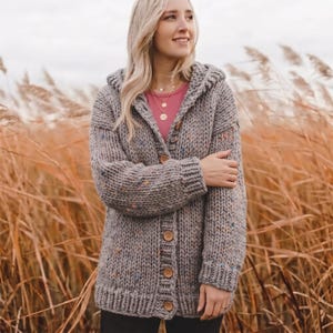 Könnte beinhalten: Eine grob gestrickte graue Strickjacke mit Kapuze, langen Ärmeln und einer Knopfleiste auf der Vorderseite mit fünf runden Holzknöpfen. Das Garn hat eine gesprenkelte Textur mit kleinen blauen, orangefarbenen und gelben Flecken im Stoff.