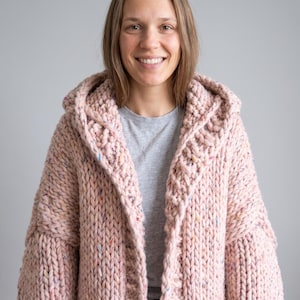 May include: A chunky knit hooded cardigan in a soft pastel pink hue, featuring a thick, textured yarn construction with subtle multicolored flecks throughout the fabric, long sleeves, and an open front design with a wide, prominent hood.