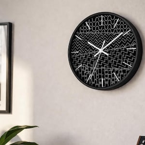 May include: A black and white wall clock with a geometric pattern. The clock has a black frame and white hands. The face of the clock features a repeating pattern of lines and shapes. The clock is mounted on a white wall.