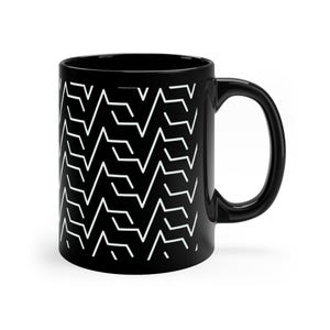 May include: Black ceramic coffee mug with a white geometric pattern. The design features a repeating zigzag pattern that covers the entire surface of the mug. The mug has a curved handle and a glossy finish.