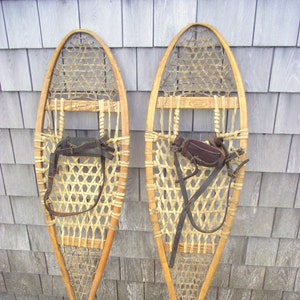 May include: Two vintage snowshoes. Each snowshoe features a wooden frame with a woven, light-colored webbing. Dark brown leather straps and bindings are attached. The snowshoes are displayed against a weathered, gray wooden background.
