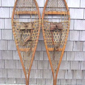 May include: Pair of vintage wooden snowshoes. Each snowshoe features a woven webbing, a wooden frame, and leather straps. The snowshoes are a light brown color and appear to be in good condition. They are designed for walking on snow.