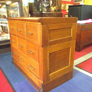 May include: A vintage wooden filing cabinet with multiple drawers and a solid wood top. The cabinet is made of a rich brown wood with a natural grain pattern. The drawers have brass pulls and are slightly recessed into the cabinet.