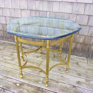 May include: A brass and glass octagonal end table with a clear glass top and a brass base with four legs. The table has a decorative design on the base.