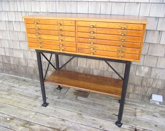 Op de afbeelding: Een houten kast met 12 lades op een zwart metalen onderstel. De kast heeft een lichte houtkleur en messing ladegrepen. Het onderstel heeft een plank onder de kast.
