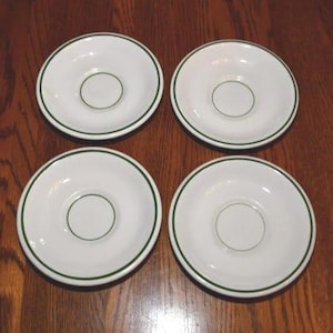 May include: Four white ceramic saucers with a green border and a green circle in the center. The saucers are arranged in a square on a wooden surface. The saucers are for use with teacups or coffee cups.