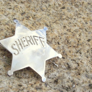 May include: A silver sheriff's star badge with the word "SHERIFF" embossed on it. The badge is lying on a rough, light brown surface.