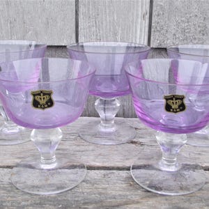 May include: Five vintage champagne glasses with a lavender bowl and clear stem and base. Each glass has a gold and black emblem. The glasses are arranged on a weathered wooden surface.
