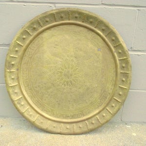 May include: A round, gold-colored metal tray with an intricate embossed design. The tray has a raised rim with a decorative border.