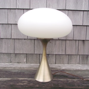 May include: A mid-century modern table lamp with a white, oval-shaped glass shade. The lamp has a brass-colored metal base with a flared, conical shape. A brown electrical cord extends from the base.