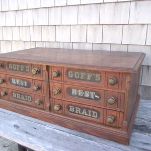 May include: Antique wooden drawer cabinet with brass knobs. The drawers are labeled "Goff's Best Braid" and "Goff's Rest Braid".