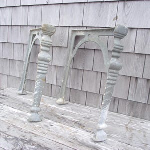 May include: Two antique cast iron table legs with a decorative design. The legs are painted a light gray color and have a weathered finish.