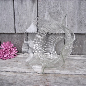 May include: A clear glass decorative bowl with a ruffled edge and textured design. A pink flower is next to the bowl on a wooden surface. The bowl has a circular center and a wavy rim.