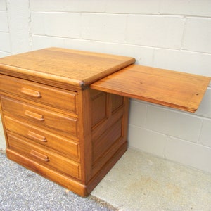 May include: A wooden chest of drawers with four drawers and a drop-down shelf. The chest is made of light brown wood and has a simple design.