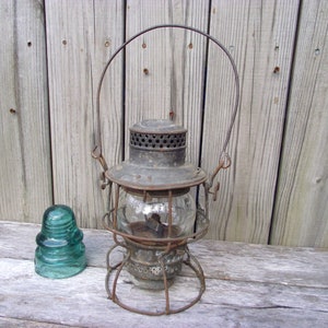 May include: An antique railroad lantern with a wire cage and a glass globe. The lantern has a metal handle and a green glass insulator next to it.
