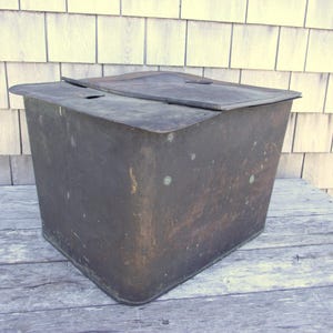 May include: A weathered, rectangular metal container with a hinged lid. The container has a dark, aged appearance, suggesting it is vintage or antique. The lid is slightly ajar, revealing a glimpse of the interior. The container is placed on a wooden surface.