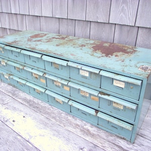 May include: A vintage light blue metal drawer cabinet with 18 drawers. The drawers have white labels with handwritten text. The cabinet has a distressed finish and shows signs of wear.