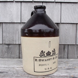May include: A vintage stoneware jug with a dark brown glazed top and handle, and a cream-colored body. The jug features a black label with the text "E. Swasey & Co. PORTL.A. ME. U.S.A." and a decorative emblem. The jug is likely used for storing liquids.