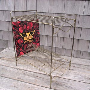 May include: A vintage gold-toned metal record rack with a top shelf and a lower section for storing vinyl records. A record with a black and red cover featuring the band Aerosmith is displayed in the rack.