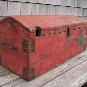 May include: A weathered red wooden toolbox with metal corner reinforcements and a handle. The toolbox has a hinged lid and a latch closure. The box is sitting on a wooden surface.