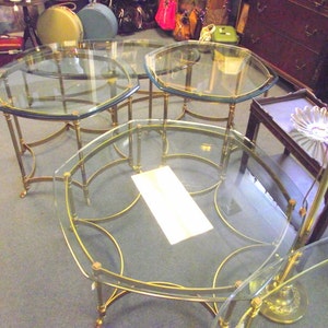 May include: Set of four glass-top tables with brass frames. The tables are nested together, with the largest table in the center and the smallest table on the right. The tables have a rounded, octagonal shape.