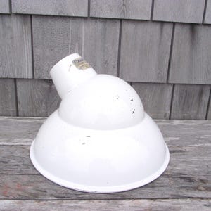 May include: A white, vintage-style industrial lamp shade with a rounded, bowl-like shape. The shade has a cylindrical top and a wider, flared bottom. The surface shows signs of wear, with small black marks.
